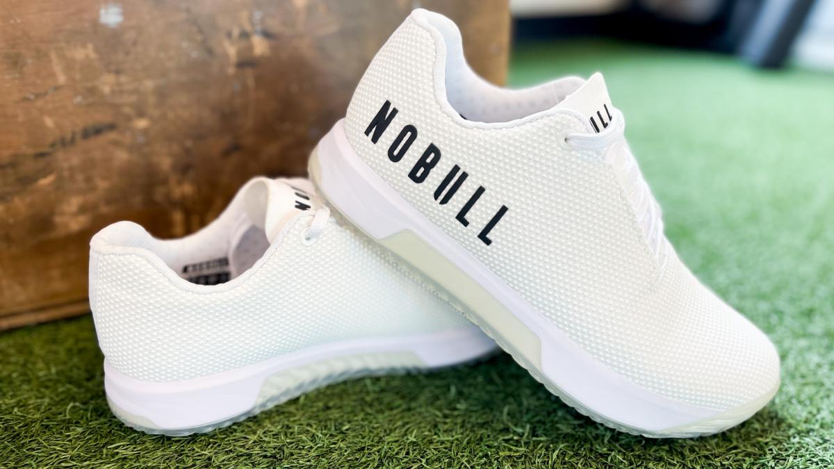 Close up photo of white Nobull Trainer+ shoes sitting on turf.
